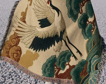 May include: A woven throw blanket featuring a large red-crowned crane in flight, surrounded by stylized green pine trees and golden-brown clouds on a cream-colored background. The blanket has a textured weave and fringed edges on the sides.