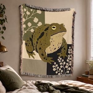 May include: A woven throw blanket featuring a large, detailed illustration of a green toad with dark spots and yellow eyes. The background is divided into rectangular blocks of olive green, cream, and black, accented with white botanical line art.