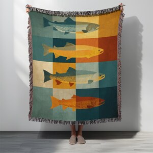 May include: A woven throw blanket featuring a repeating pattern of colorful fish illustrations. The blanket has a fringed edge and is displayed against a white background. The fish are in shades of blue, yellow, and orange, set against a grid of complementary colors.