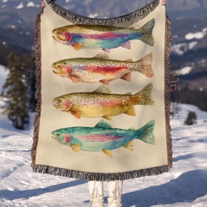 Könnte beinhalten: Eine beige Decke mit vier Aquarellillustrationen von Forellen. Die Fische sind in verschiedenen Farben dargestellt, darunter Blau, Gelb und Rot, mit detaillierten Schuppen und Flossen. Die Decke hat einen Fransenrand.