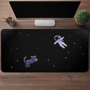 Puede incluir: Alfombrilla de escritorio rectangular negra con un diseño pixelado de un gato y un astronauta flotando en el espacio con estrellas. La alfombrilla está colocada sobre un escritorio de madera, con un monitor de ordenador, una pequeña planta y un portalápices en el fondo.