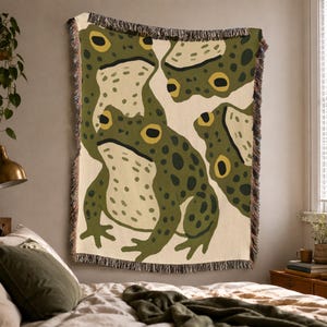 May include: A woven throw blanket featuring a repeating pattern of green frogs with dark spots and yellow eyes on a cream background. The blanket has a textured surface and frayed edges along the top and bottom, showcasing a detailed textile design.