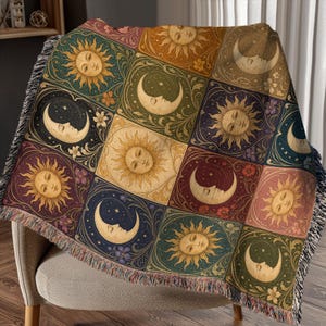 May include: A woven throw blanket with a repeating pattern of suns and crescent moons with faces, in gold, blue, and burgundy. The blanket has a fringed edge and is draped over a chair. The blanket is made of a soft material.