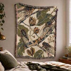 May include: A woven throw blanket featuring a repeating botanical and insect pattern. The design displays detailed illustrations of cicadas perched on tree branches with dark green leaves and brown seed pods against a cream-colored background.
