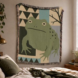 May include: A woven throw blanket featuring a large green frog centered against a background of geometric shapes, including black triangles and light blue vertical stripes, with a dark branch silhouette and a decorative fringed edge on all sides.
