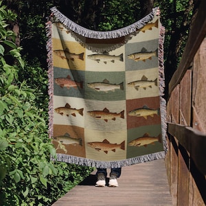Peut inclure: Une couverture tissée avec un motif répété de poissons de différentes couleurs. La couverture a une bordure à franges et est présentée à l'extérieur. Les poissons sont représentés dans des tons de marron, de beige et de rouge, sur un fond de carrés aux tons sourds.