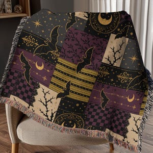 May include: A decorative throw blanket with a patchwork design featuring bats, moons, stars, and ornate floral patterns in black, gold, purple, and beige. The blanket has a fringed edge and is draped over a chair.