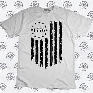 May include: A white t-shirt featuring a black distressed American flag design. The flag has 13 stripes and a circle of stars with the year 1776 in the centre.