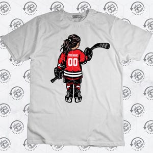 K&ouml;nnte beinhalten: Ein wei&szlig;es T-Shirt mit einem roten und schwarzen Hockey-Spieler-Motiv. Der Spieler ist ein M&auml;dchen, das ein Trikot mit dem Text "YOUR NAME" und der Nummer "00" tr&auml;gt.