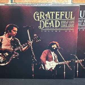 May include: Two vinyl record covers for Grateful Dead's "The Wharf Rats Come East" album. The covers are red and black with the band's name and album title in white. The first cover is labeled "Volume One" and features a photo of Jerry Garcia playing a guitar. The second cover is labeled "Volume Two" and features a photo of the band.