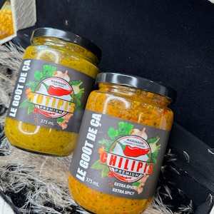 Peut inclure: Deux pots en verre de marinade avec des couvercles noirs. L'un est étiqueté "TUTIPIS PREMIUM" et l'autre "CHILIPIS PREMIUM". Les deux pots portent des étiquettes avec du texte en français, dont "LE GOÛT DE ÇA" et "375 ml". Le pot "CHILIPIS" est étiqueté "EXTRA ÉPICE".