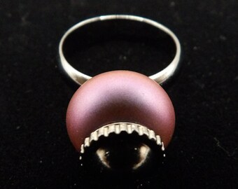Puede incluir: Un anillo de plata con una gran piedra preciosa redonda e iridiscente de color púrpura. La piedra preciosa tiene un borde texturizado y festoneado.