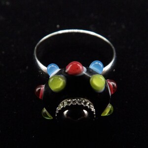 May include: A silver ring with a black glass bead set with colorful glass accents. The bead has a textured surface and is surrounded by a silver band.