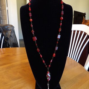May include: A red beaded lanyard with a red and green floral charm. The lanyard is made of small square beads and round beads.