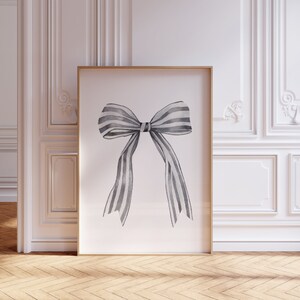 May include: A framed art print featuring a watercolor illustration of a bow with gray and white stripes. The artwork is displayed in a gold frame and leans against a white wall with decorative molding, set on a wooden floor.