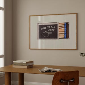 May include: A framed artwork featuring a matchbox with matches and the words "DRAMATIC EXIT" on a chalkboard. The matches have blue tips. The artwork is in a wooden frame and hangs on a neutral-colored wall.