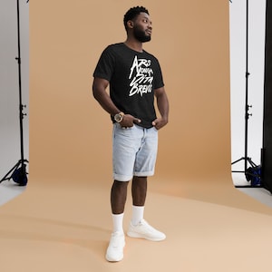Peut inclure: Une personne portant un t-shirt noir avec du texte blanc qui dit "ARS LONGA VITA BREVIS". La personne porte un short en jean bleu et des baskets blancs. L'arrière-plan est de couleur beige clair.