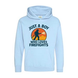 Può includere: Felpa con cappuccio azzurra con la scritta "JUST A BOY WHO LOVES FIREFIGHTS". La grafica presenta la sagoma di un pompiere con un tubo contro uno sfondo di tramonto arancione e giallo. Un top comodo e casual.