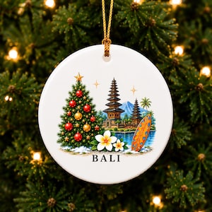Puede incluir: Un adorno redondo de cerámica blanca que cuelga de un cordón dorado. El diseño muestra un árbol de Navidad decorado, un templo balinés, una tabla de surf y flores de plumeria blancas. La palabra BALI está impresa en negro en la parte inferior.
