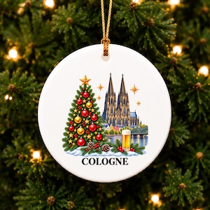 Könnte beinhalten: Ein runder, weißer Keramik-Weihnachtsbaumschmuck mit einer farbigen Illustration eines Weihnachtsbaums mit roten und goldenen Kugeln, dem Kölner Dom, einem Glas Bier und dem Schriftzug COLOGNE. Der Anhänger hängt an einer goldenen Schnur.