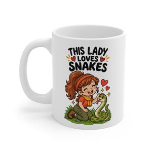 Puede incluir: Una taza de cerámica blanca con una ilustración de dibujos animados de una persona de cabello castaño arrodillada en la hierba verde, acariciando una serpiente verde. El texto "THIS LADY LOVES SNAKES" está impreso en negro sobre la ilustración, con corazones rojos.