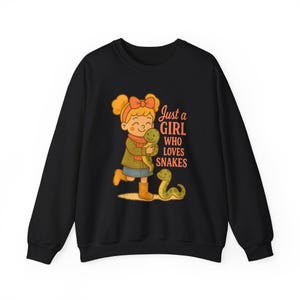 May include: Black sweatshirt with a cartoon illustration of a girl with orange hair and a bow, hugging a green snake. The text "Just a GIRL WHO LOVES SNAKES" is printed on the front, with another green snake.