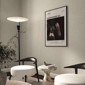 May include: A black and white poster of Muhammad Ali boxing with the text "Muhammad Ali" and "55-5-0" on it. The poster is hanging on a white wall in a living room with two chairs and a side table with a book on it.