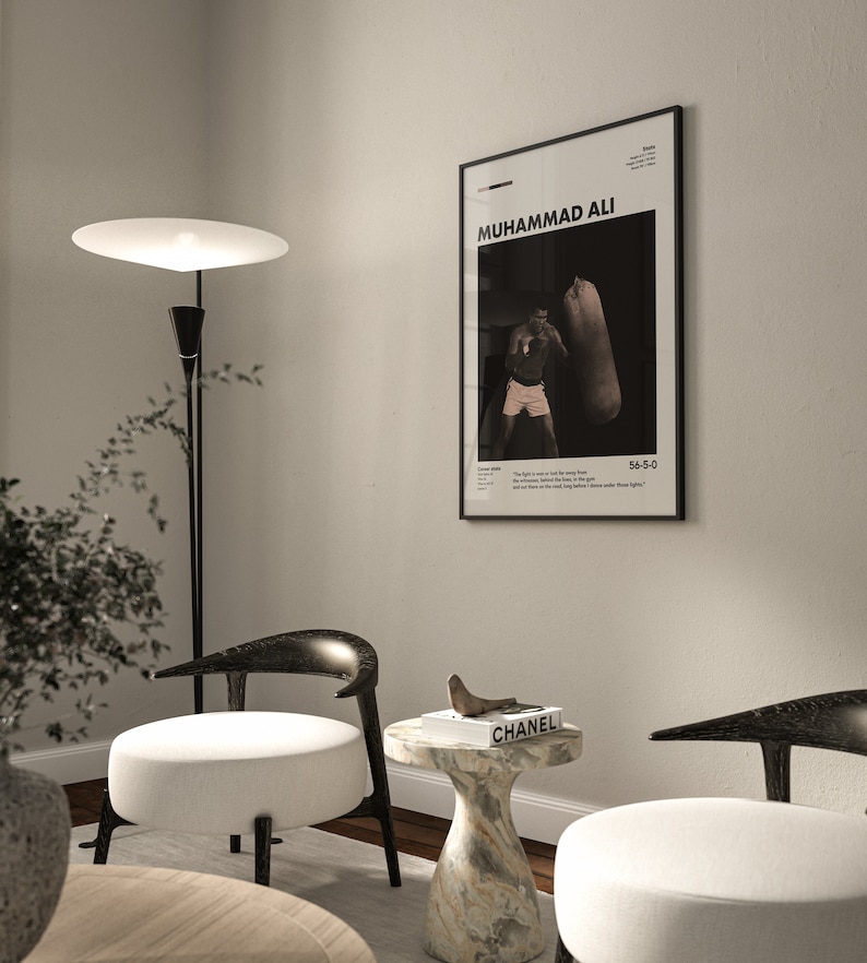 May include: A black and white poster of Muhammad Ali boxing with the text "Muhammad Ali" and "55-5-0" on it. The poster is hanging on a white wall in a living room with two chairs and a side table with a book on it.