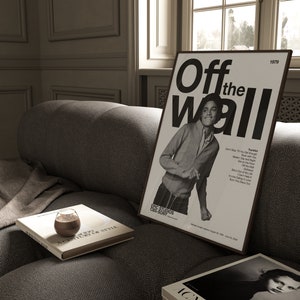 May include: A black and white poster with the text "Off the Wall" and a photo of Michael Jackson. The poster is framed and hanging on a wall in a room with a couch and a window.