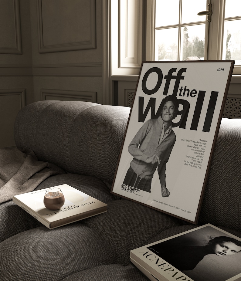 May include: A black and white poster with the text "Off the Wall" and a photo of Michael Jackson. The poster is framed and hanging on a wall in a room with a couch and a window.