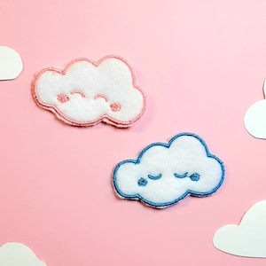 May include: Two felt clouds with embroidered faces on a pink background. One cloud is pink with a smiling face and the other is blue with a sleepy face.