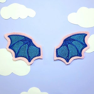 May include: Two embroidered blue dragon wing patches with pink felt edging, set against a light purple background with white paper cloud cutouts. The wings have detailed stitching and a symmetrical design.