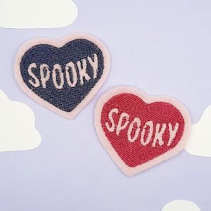 May include: Two heart-shaped patches with the word "SPOOKY" embroidered on them. One patch is navy blue with a light pink border, and the other is red with a light pink border. The background is a light purple with white cloud cutouts.