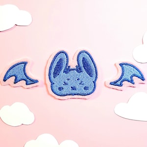 May include: A blue embroidered bat patch with a cute face and bat wings. The patch is surrounded by a pink border. The background is a light pink color with white cloud cutouts.