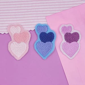 May include: Three heart-shaped embroidered patches in pink, blue, and purple. Each patch features a layered design with varying shades of the same color, outlined with a contrasting color. The patches are on a pink and purple background.