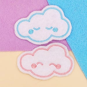 May include: Two white cloud-shaped felt appliques with embroidered details. One cloud has blue stitching and accents, the other has pink. Both have smiling faces and are set against a purple and blue background.