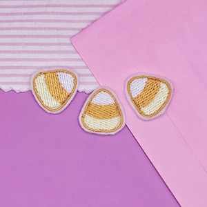 May include: Three candy corn-shaped embroidered patches. Each patch features a white, yellow, and orange color scheme with a stitched border. The patches are arranged on a purple and pink background, with a striped fabric in the upper left corner.