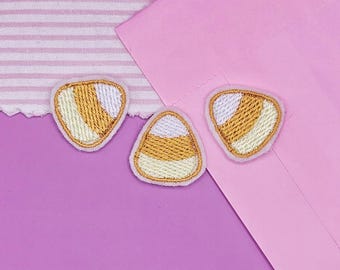 May include: Three candy corn-shaped embroidered patches. Each patch features a white, yellow, and orange color scheme with a stitched border. The patches are arranged on a purple and pink background, with a striped fabric in the upper left corner.