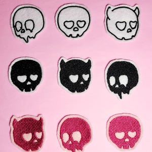 May include: Nine embroidered iron-on patches featuring skull designs in white, black, and pink. The skulls have hearts for eyes and some have devil horns.