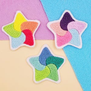 May include: Three colorful star-shaped embroidered patches. Each star features a different arrangement of rainbow colors, including red, orange, yellow, green, blue, and purple. The patches have a white border and are set against a pastel background.