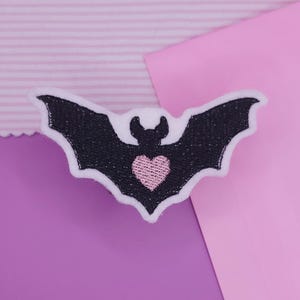 May include: A black bat-shaped patch with white trim and a pink heart in the center. The bat has outstretched wings and is set against a background of pink and purple paper.