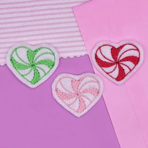 May include: Three heart-shaped embroidered patches with a candy cane design. One is green and white, one is pink and white, and the third is red and white. The patches are on a purple surface with pink and striped fabric.