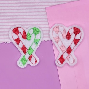 May include: Two heart-shaped patches with candy cane designs. One features red and green candy canes, the other has pink and red candy canes. The patches are white with embroidered details, perfect for holiday crafts.