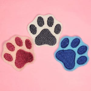May include: Three embroidered paw print patches in different colors: pink, blue, and white. The patches are on a pink background.