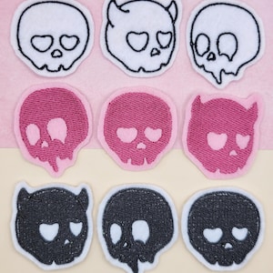 May include: Nine embroidered skull patches in three colors: white, pink, and black. Each skull has heart-shaped eyes and a white border. Some skulls have horns, and others have a dripping effect. The patches are arranged on a pink and beige background.