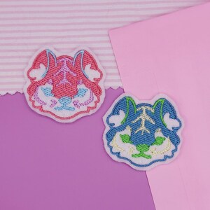 May include: Two embroidered patches in the shape of a stylized tiger face. One is pink and red, the other is blue and green. Both have white borders and are on a pink and purple background.
