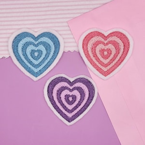May include: Three heart-shaped embroidered patches in shades of blue, pink, and purple. Each patch features concentric heart designs and a white border. The patches are arranged on a pink and purple background.