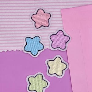 May include: Five colorful star-shaped patches. Each patch has a different pastel color: pink, blue, purple, and yellow-green. The stars have a white border and black outline. The patches are on a pink and white striped fabric and a solid pink background.
