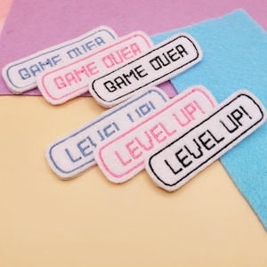 May include: Six rectangular white fabric patches with rounded corners and embroidered borders. Three patches feature the text GAME OVER in blue, pink, and black. Three patches feature the text LEVEL UP! in blue, pink, and black thread colors.