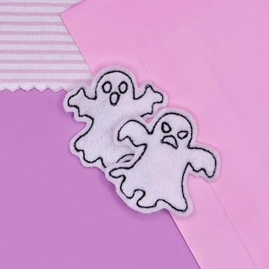 May include: Two white felt ghost-shaped appliques with black outlines and facial features. One ghost has an open mouth, and the other has a frowning expression. The appliques are on a pink and purple background.